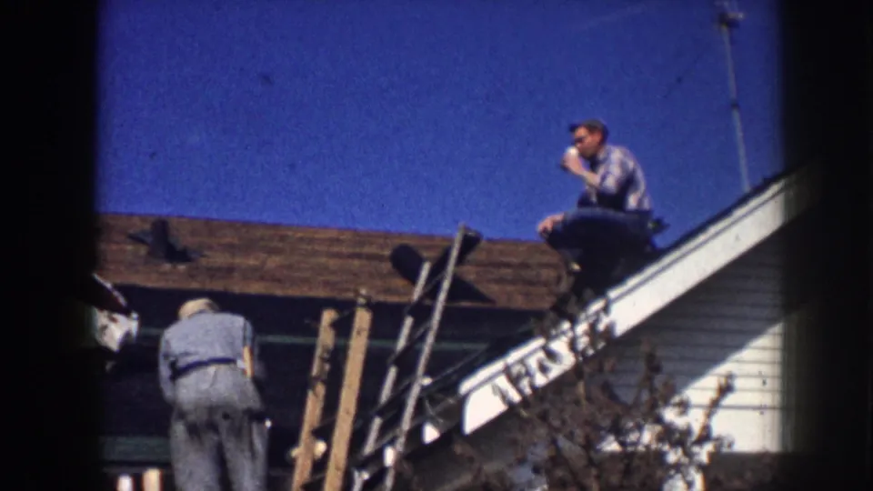 1958 Nebraska home-movie frame of two men on a house roof, with one seated and drinking while another works lower on the roof.