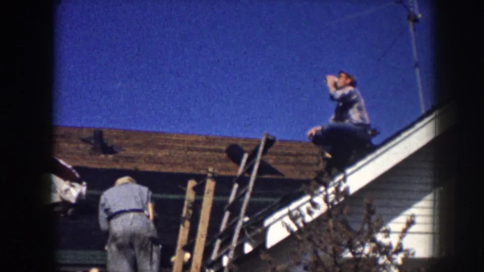 Men on a Nebraska house roof in 1958, one taking a drink from a cup as another works below beside ladders and roofing materials.