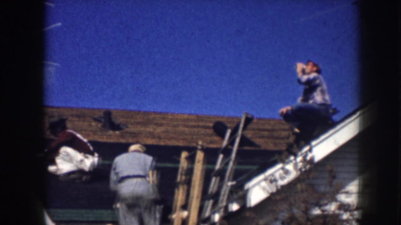 Men working on a shingled roof beside a white house under a blue sky.