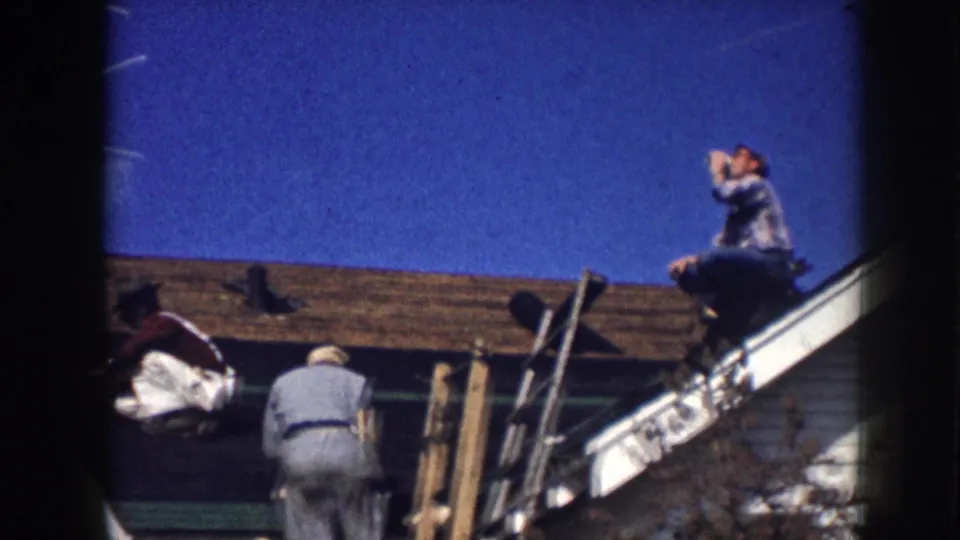 1958 Nebraska 8mm frame showing men on and near a residential roof, with white siding and green trim visible.