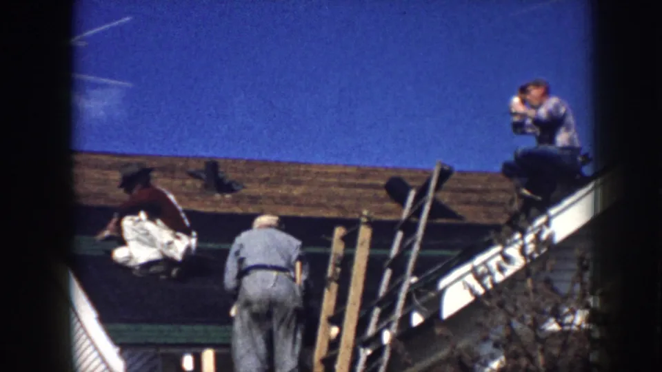 1958 Nebraska home-movie frame with a kneeling man on a roof and another man standing near the house edge.