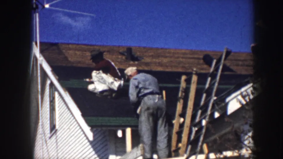 1958 Nebraska 8mm frame showing roof repair activity on a white house, with one man standing and another on the shingles.