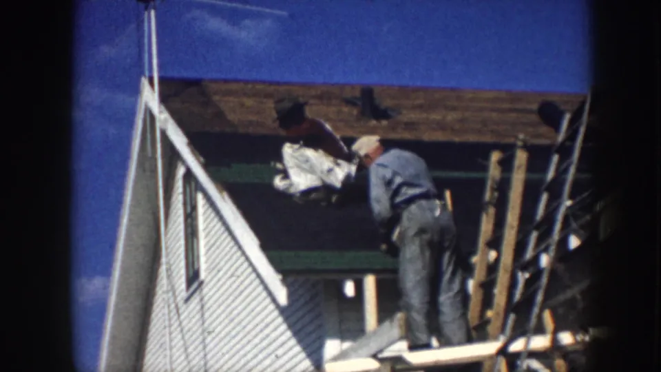1958 Nebraska home-movie frame of men handling roof work near white siding, green trim, and leaning boards.