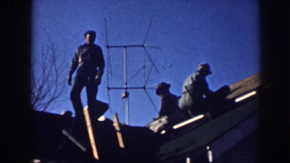 Men work and stand on a Nebraska house roof in 1958, with an antenna and roof edge visible against the sky.