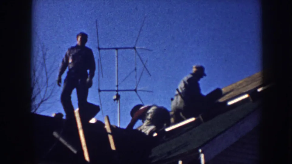 A 1958 Nebraska frame shows men bent over and standing on a sloped house roof near a rooftop antenna.
