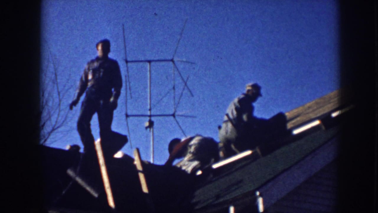 1958 Nebraska home-movie frame showing men working on a residential roof beside a large outdoor TV antenna.