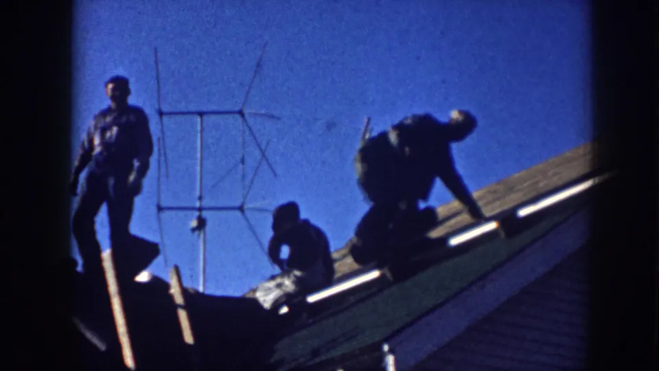 Backlit 1958 Nebraska roof-work scene with men crouching and leaning on a sloped residential roof near an antenna.
