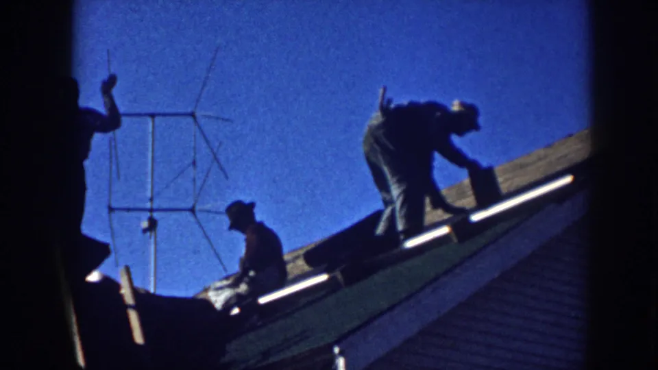 Late-1950s Nebraska home-movie frame with silhouetted men on a residential roof, one gesturing while others handle roofing material.
