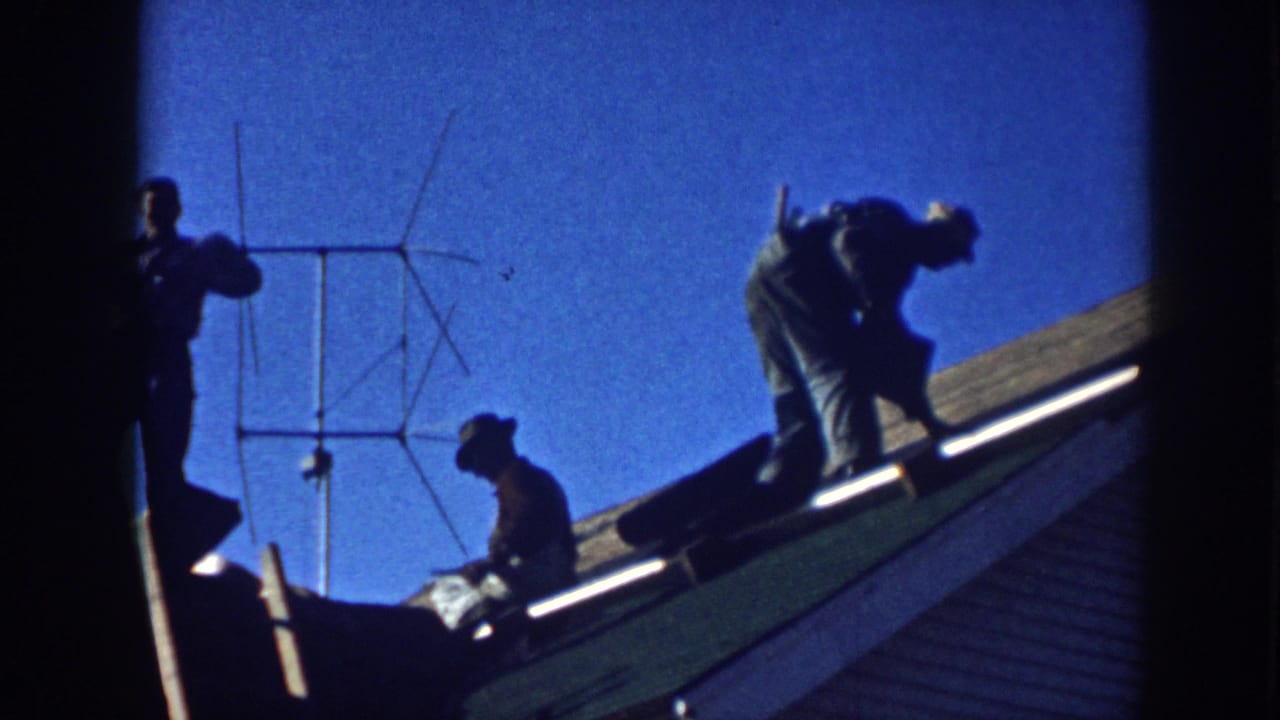 Men work on a sloped house roof near a TV antenna under a blue sky.