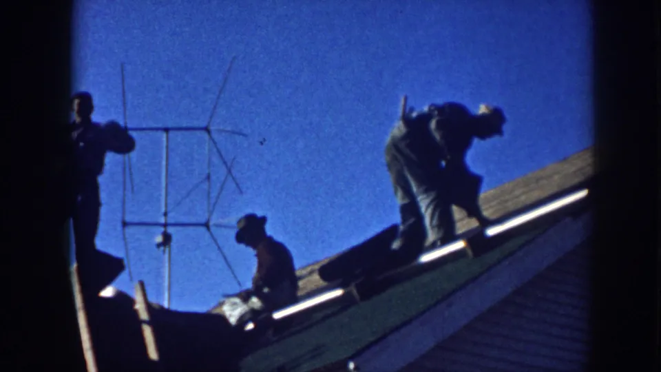 In Nebraska 1958 home-movie footage, several men appear on a sloped house roof with roofing material and a TV antenna nearby.