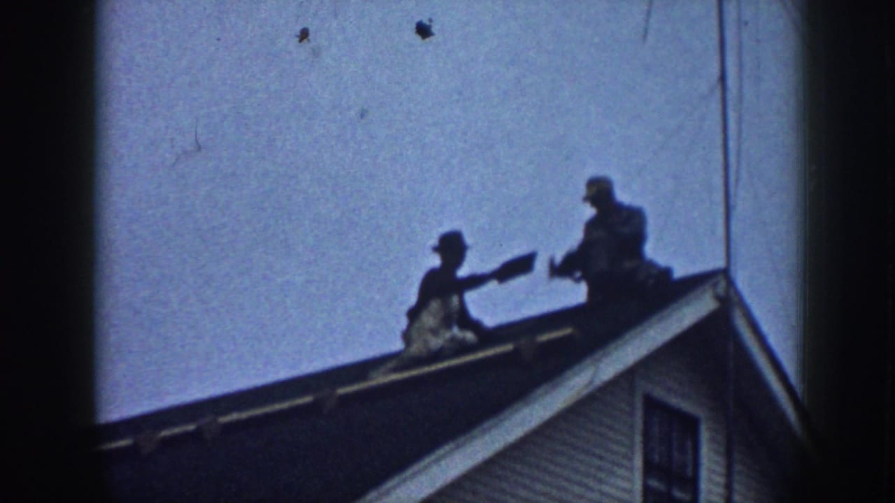 Two figures sit or kneel on a roof ridge while handling roofing material.