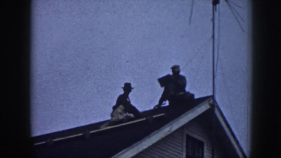 The roof repair continues with two figures on the sloped roof of the house, one lower on the slope and one higher near the antenna line.