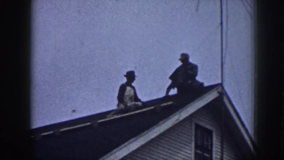 Two men in hats appear on a Nebraska house roof in 1958 home-movie footage, viewed from below against the sky.