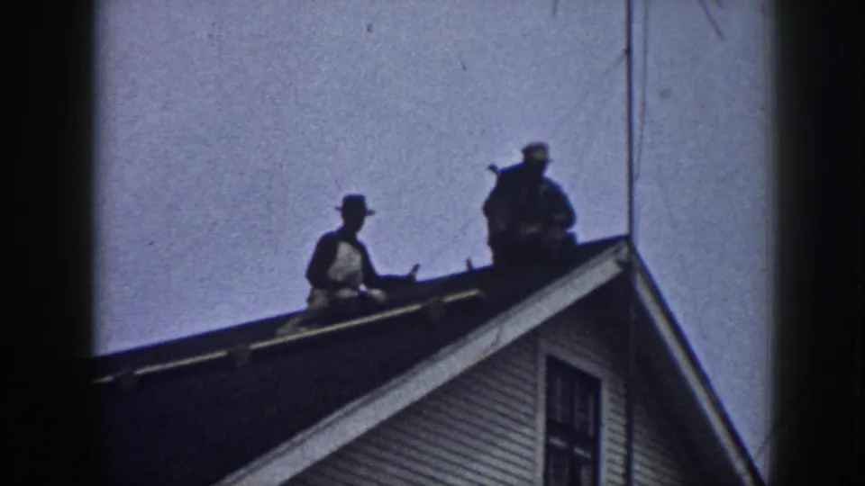 Two hat-wearing men sit on a sloped roof in 1958 Nebraska home-movie footage, with white siding below and sky behind them.