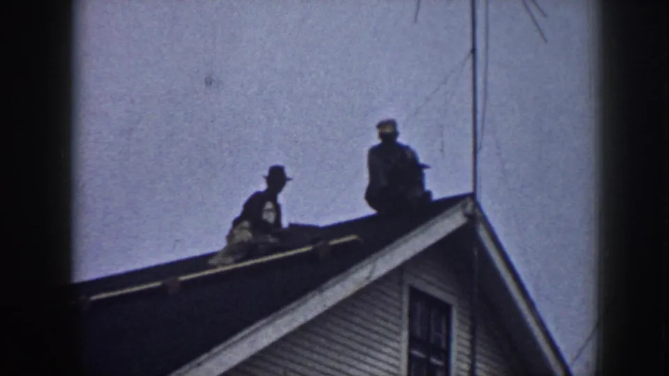 A low-angle 1958 Nebraska home-movie frame shows two men on a roof, silhouetted above white clapboard siding and a window.