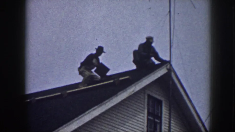 In a 1958 Nebraska roof-repair scene, one man bends toward the shingles while another sits near the edge of the white house roof.