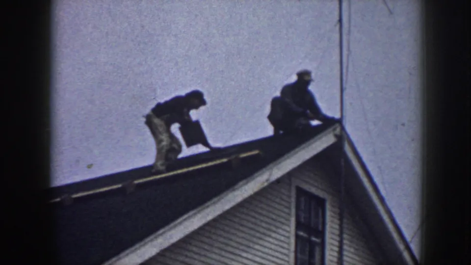 1958 Nebraska 8mm frame showing two men working on a house roof under a pale sky.