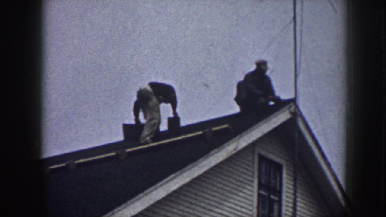 One man bends over roofing material while another sits near the roof ridge.