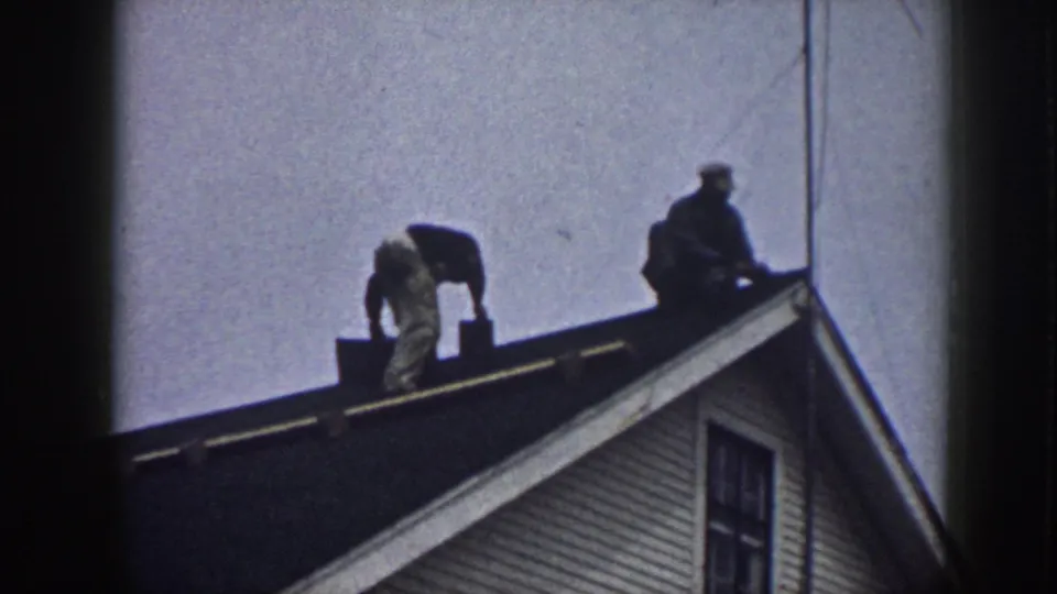 Two adult men appear on a Nebraska house roof during shingle work in a soft 8mm frame.