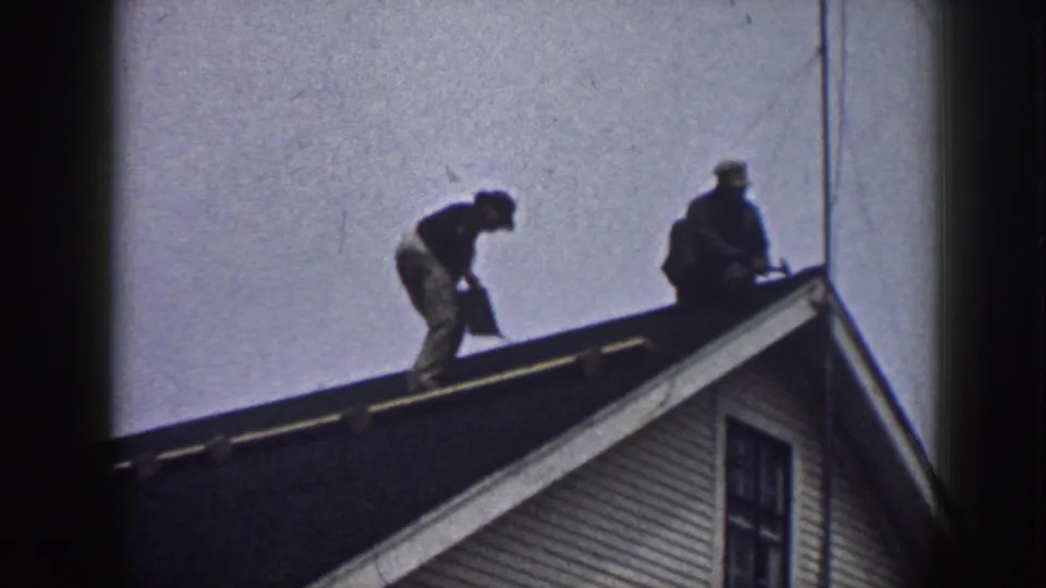 1958 home-movie frame of two men continuing roof repair on a Nebraska house exterior.