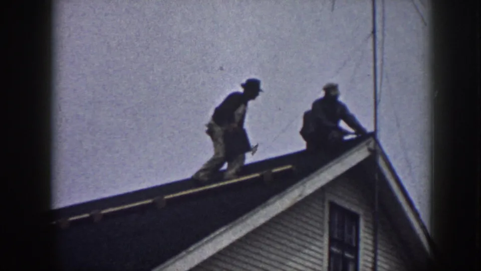 Nebraska 1958 8mm still showing two men on a residential roof during shingle replacement.