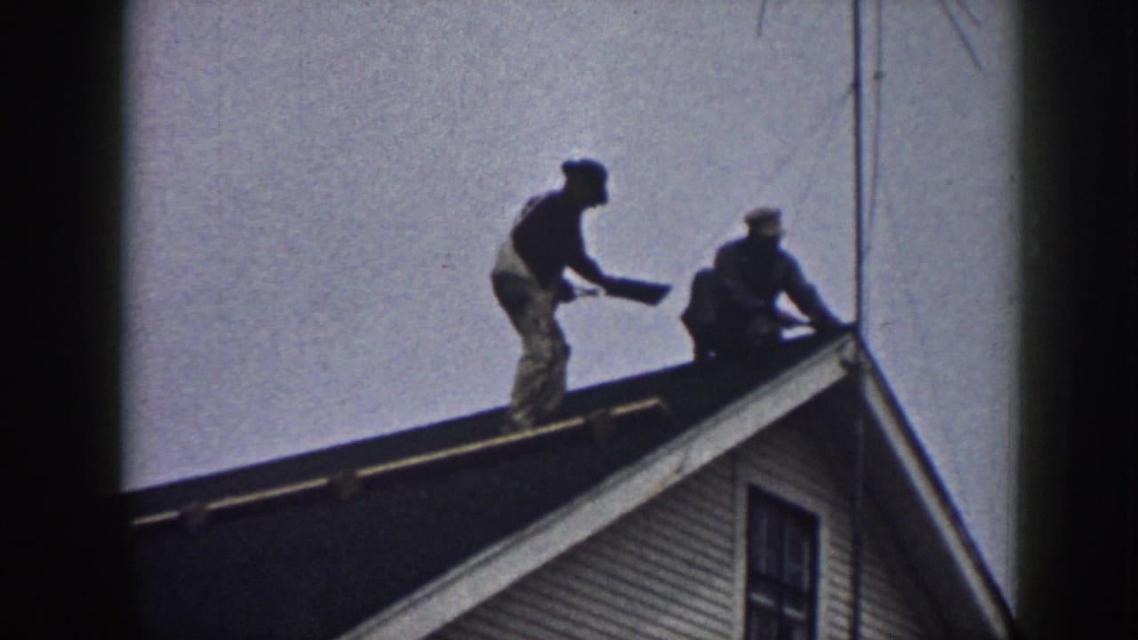 The standing worker holds a flat piece of material toward the crouching worker on a Nebraska residential roof in the 1958 home movie.