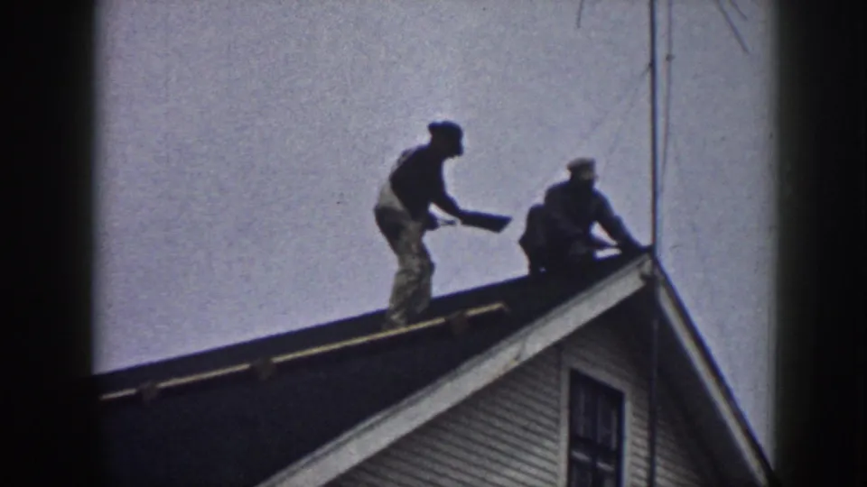 The standing worker holds a flat piece of material toward the crouching worker on a Nebraska residential roof in the 1958 home movie.