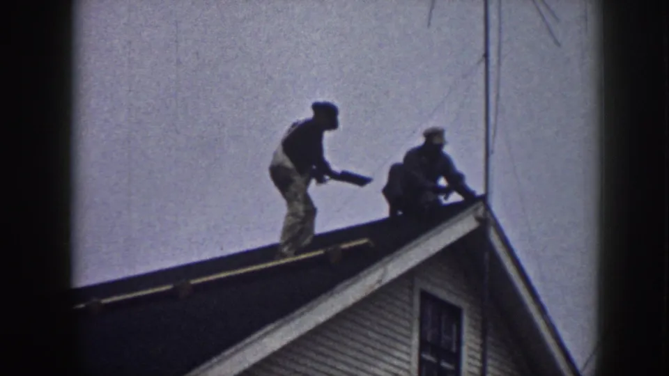 Two men remain on the sloped roof, with one bending forward and the other crouched near the edge of the visible house exterior.