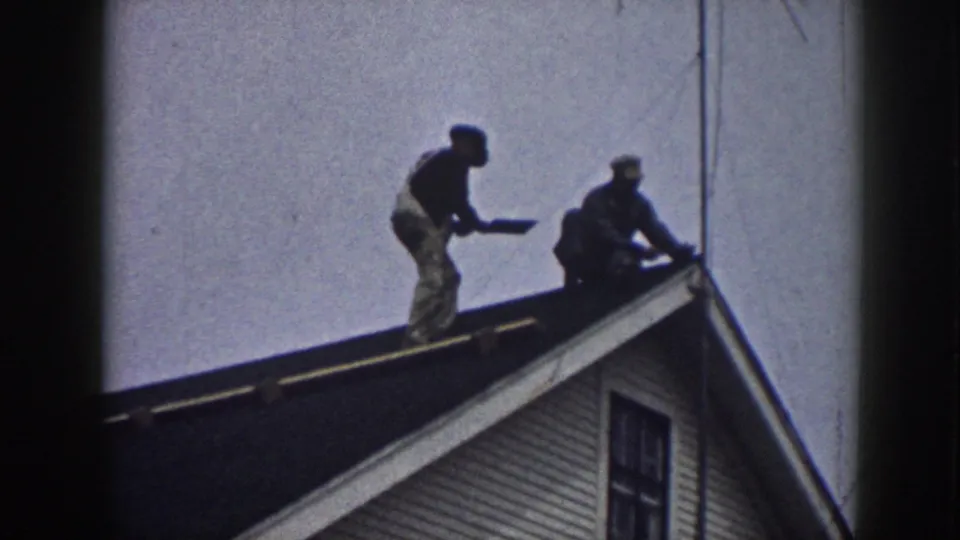 Near the end of the clip, one man stands holding roofing material while the other stays crouched beside the roof ridge on the Nebraska house.