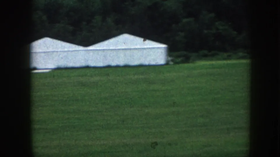 1958 Nebraska rural view of a long white-roofed structure at the edge of a green field, backed by trees.
