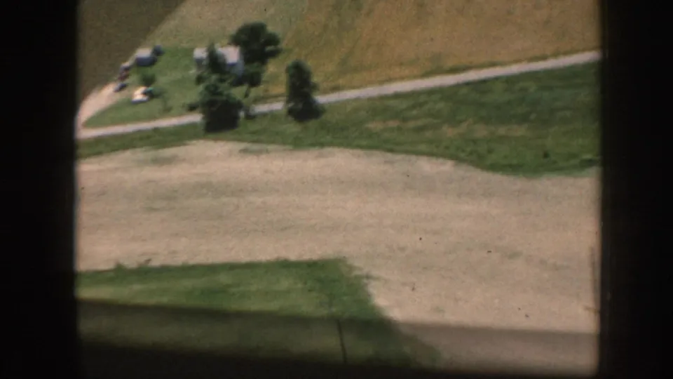 Elevated 1958 Nebraska 8mm view of a dirt or gravel path, open fields, trees, and a small distant building cluster.