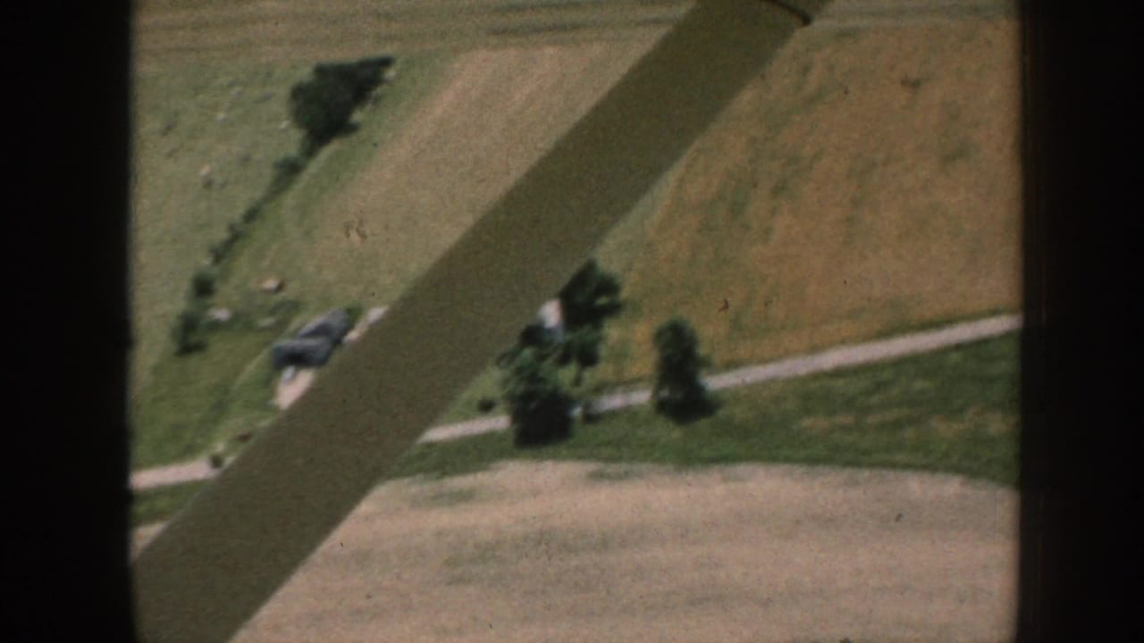 1958 Nebraska aerial home-movie view from a small aircraft over fields, trees, farm roads, and gray rural buildings.