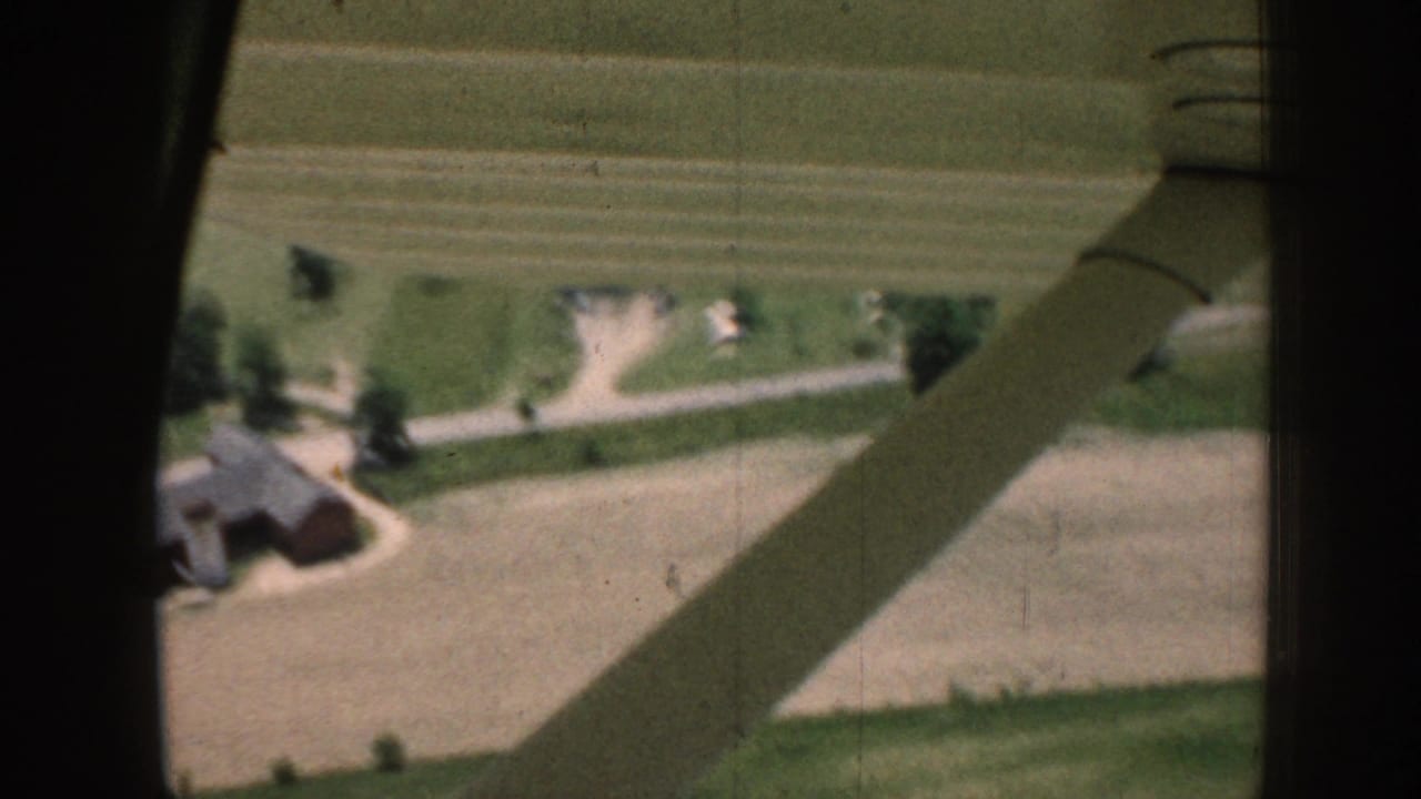 Wing strut crossing an aerial view of fields, trees, roads, and farm buildings.