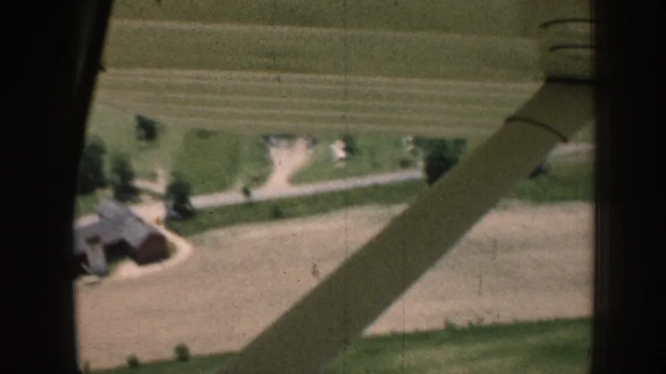Aerial 1958 Nebraska view with aircraft struts framing farm roads, cultivated fields, trees, and gray outbuildings below.