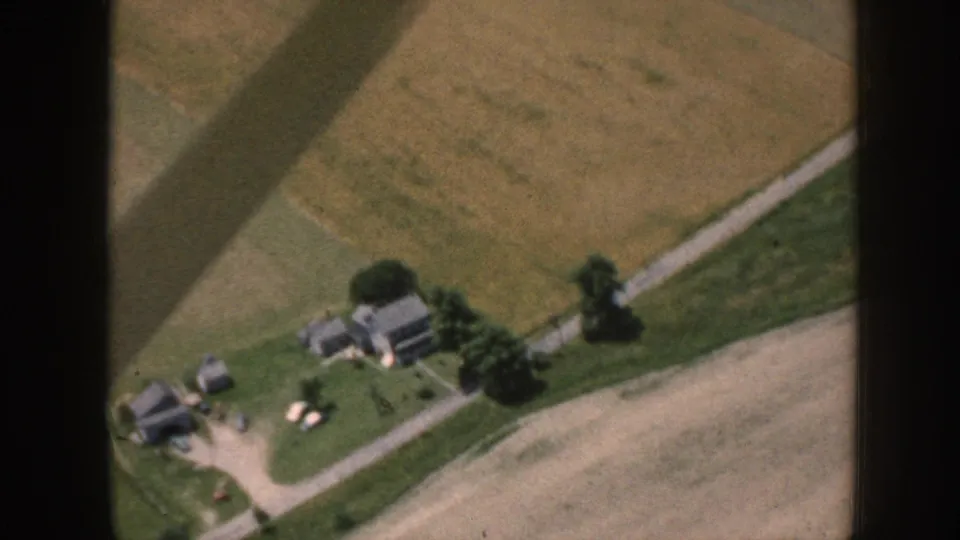1958 Nebraska aerial footage shows a farmstead, outbuildings, green tree cover, roads, and tan fields from a small aircraft.