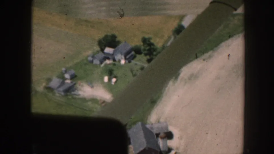 Aerial home-movie frame from 1958 Nebraska with aircraft struts over a farm cluster, trees, road, and pale cultivated field.