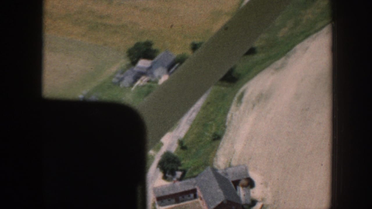 Aerial view of rural buildings, fields, trees, and a dirt road through a dark film-frame border.
