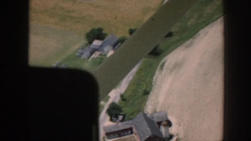 1958 Nebraska aerial 8mm frame showing farm buildings, green fields, trees, and a pale dirt road from above.