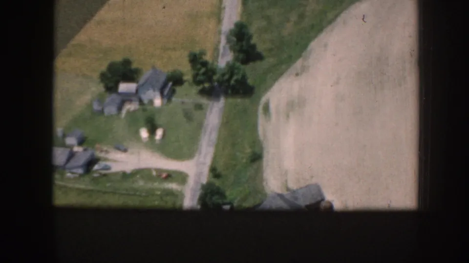 1958 Nebraska aerial frame with scattered farm structures, a road, green grass, trees, and adjacent cultivated fields.