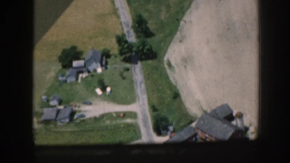 1958 Nebraska aerial 8mm frame showing rural buildings near a road, with fields and trees surrounding the property.