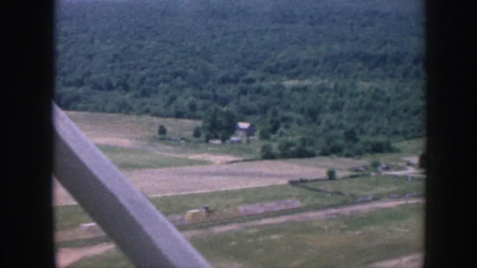 1958 Nebraska aircraft-window view of farmland, a small building cluster, roads, and wooded terrain below.