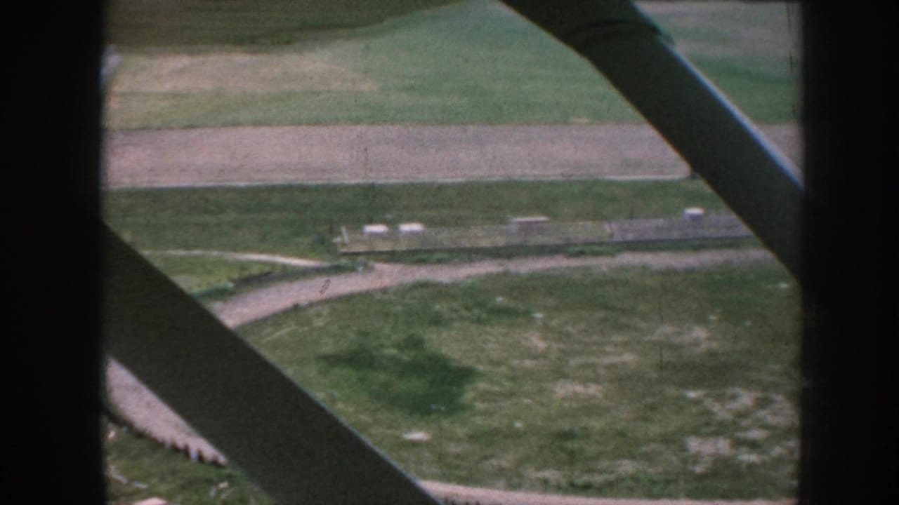 Airplane window struts over fields, dirt paths, and a small rural airfield area.