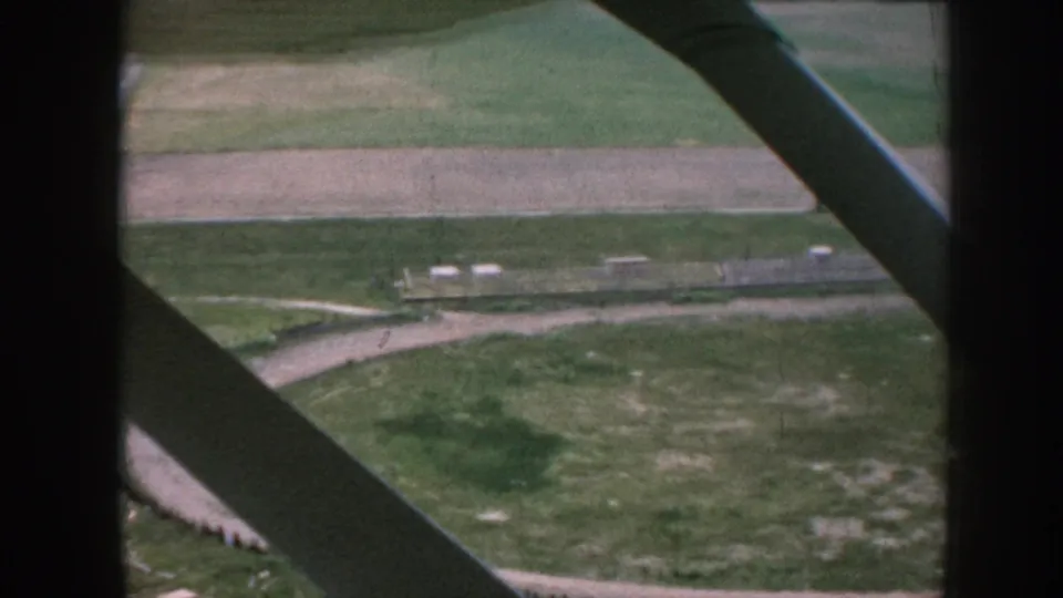 1958 Nebraska home-movie view through an airplane window, looking down at fields, paths, and a small rural airfield area.