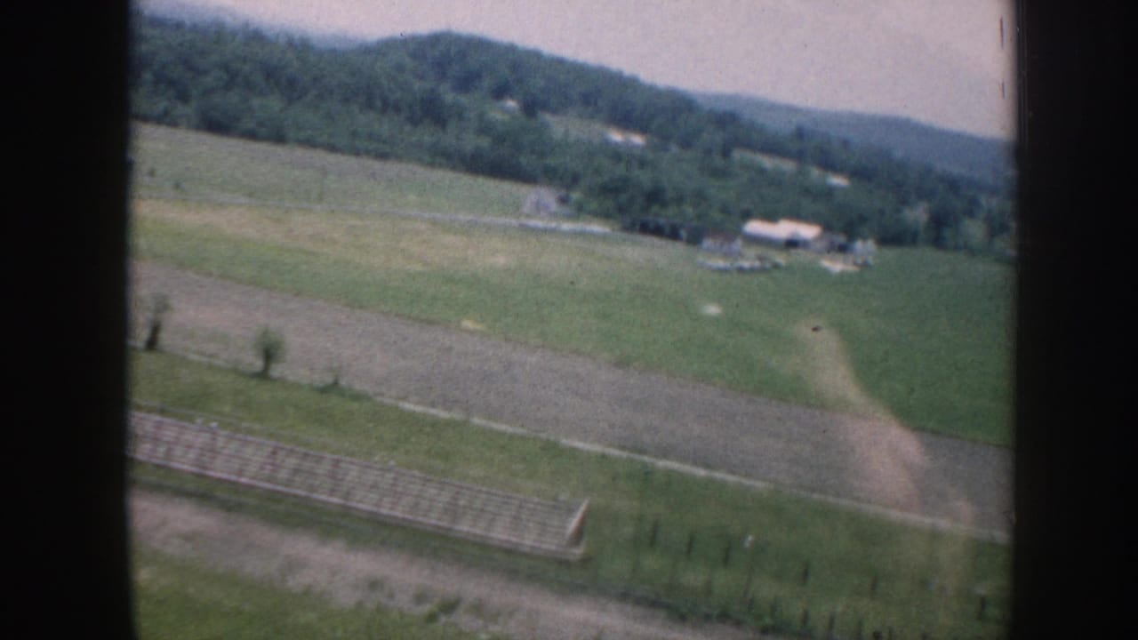 Aerial 1958 Nebraska view of green farmland, low wooded hills, scattered buildings, and a runway-like strip below.