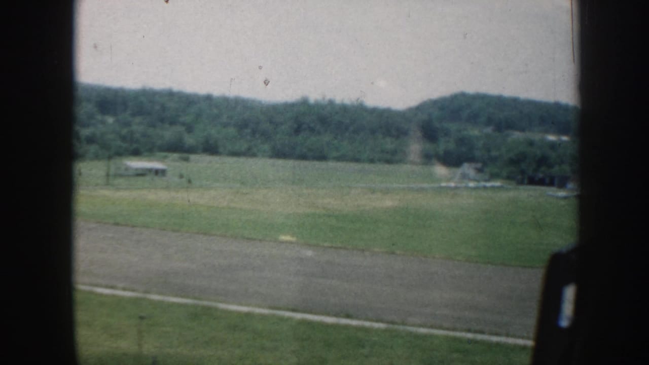 Green field and wooded hills seen through a car window with a dirt road below.