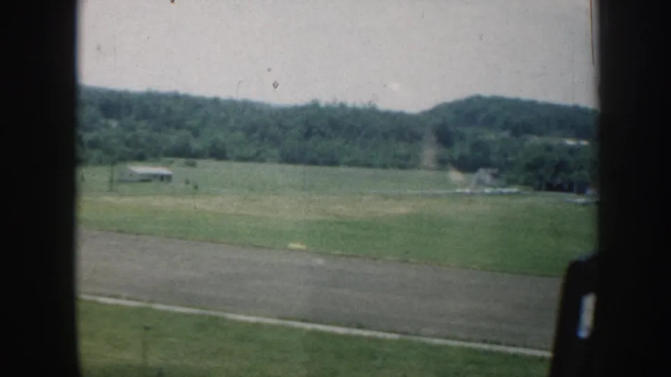 1958 Nebraska rural field and wooded hills filmed from inside a moving car, with window frame and road visible.