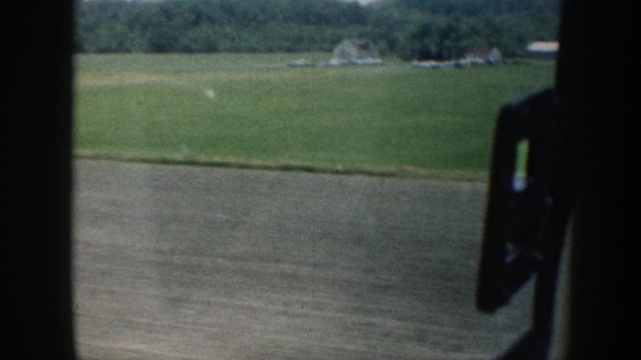 A 1958 Nebraska countryside view through a car window, showing fields, distant farm structures, and wooded hills.