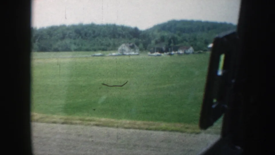 Nebraska farmland in 1958 appears beyond the car window, with a green field, small buildings, wooded hills, and visible film wear.