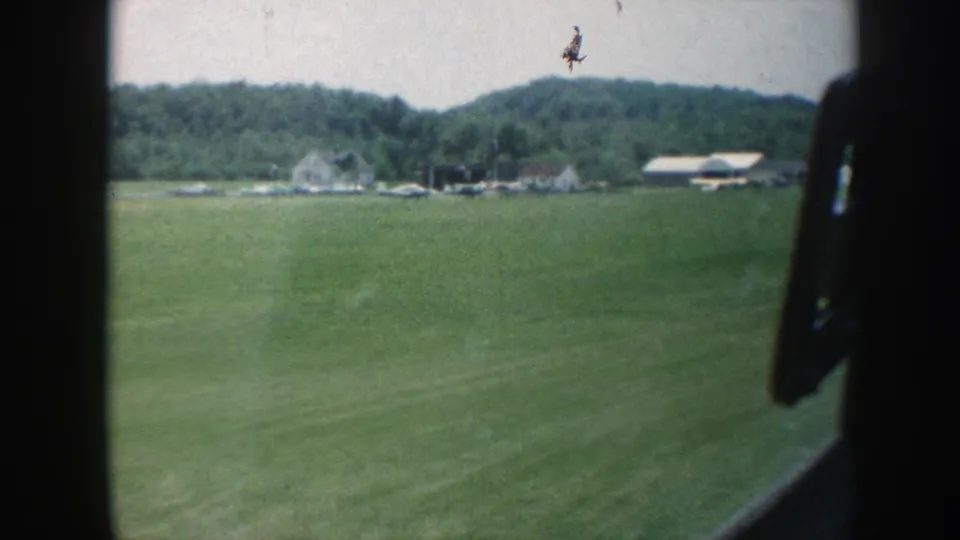 A late-1950s Nebraska home-movie frame shows open farmland and distant farm buildings through a vehicle window.