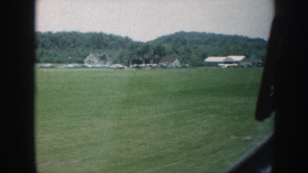 1958 Nebraska home-movie frame showing a green rural field, distant pale buildings, trees, and dark foreground framing from a moving viewpoint.